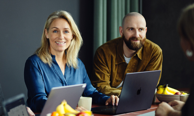 To mennesker sidder ved et bord med computere foran sig, og den ene person kigger ind i kameraet og smiler