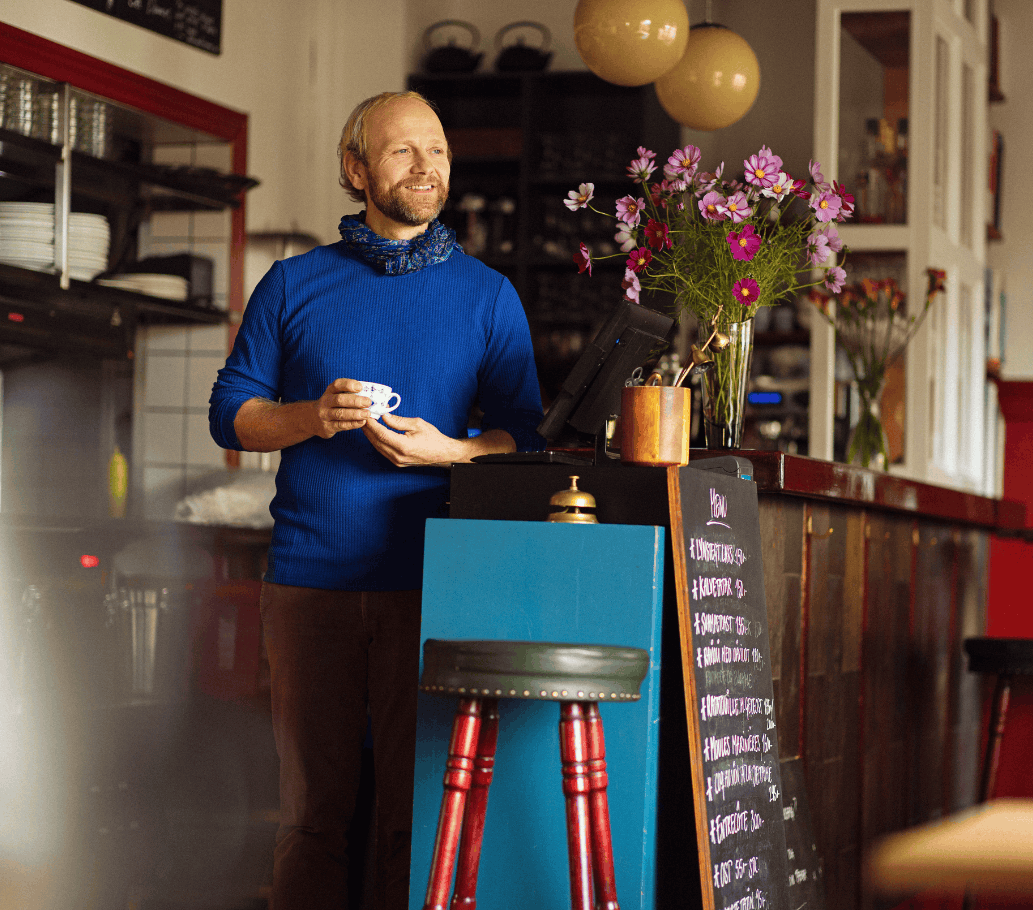 En mand står med en kop kaffe i hånden i en restaurant og smiler