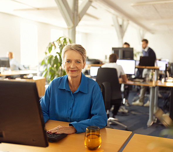 En kvinde sidder ved sit skrivebord foran en computer og smiler