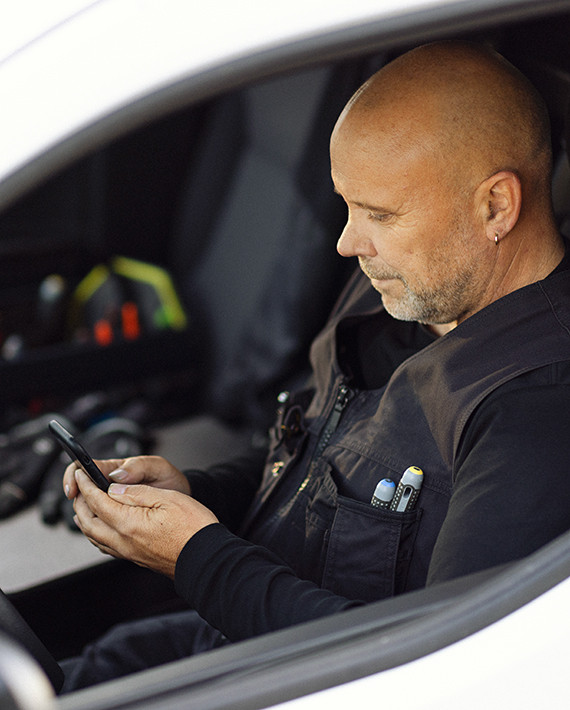 En mand sidder i sin bil fyldt med værktøj og kigger på sin mobiltelefon