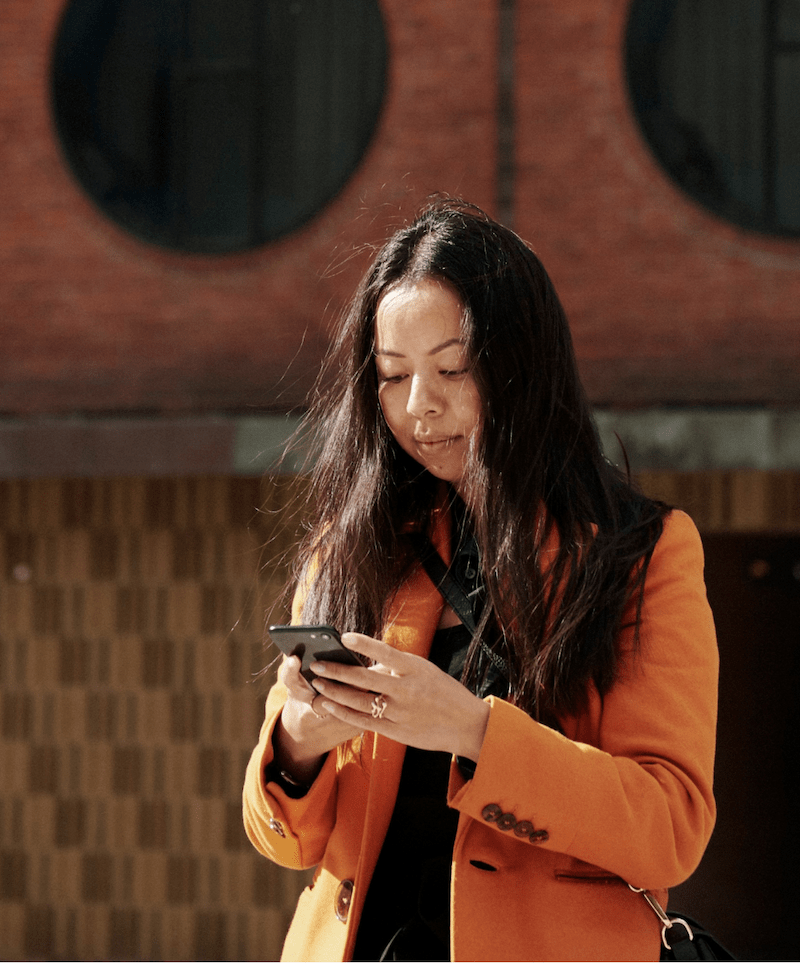 En kvinde med en orange jakke på står og kigger på sin mobiltelefon