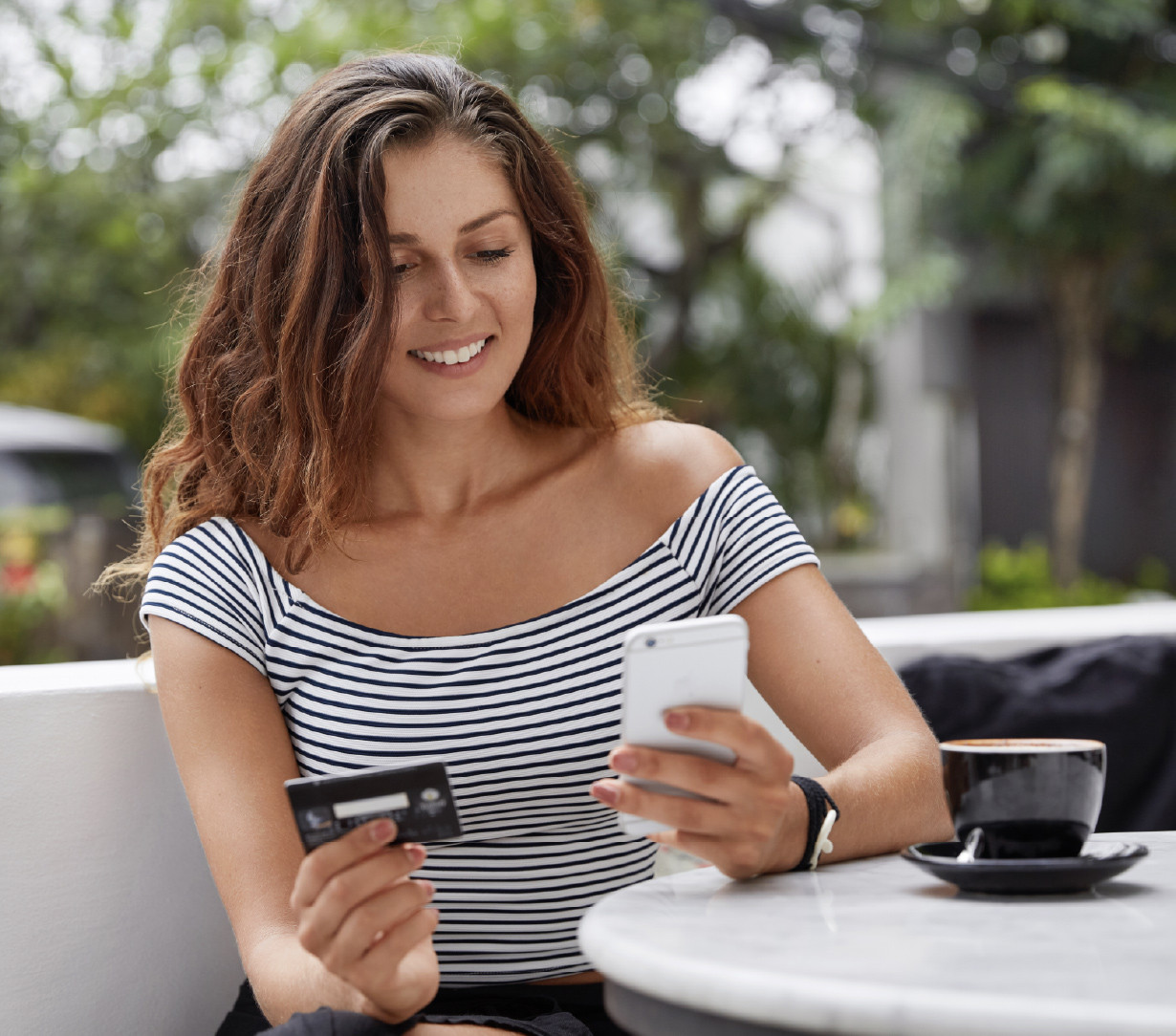En kvinde sidder og smiler, mens hun kigger på sin smartphone og et betalingskort