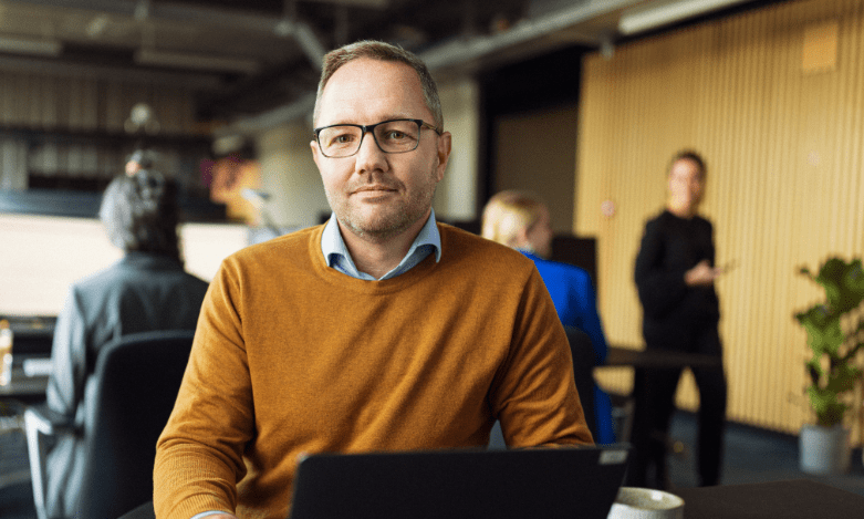 En mand med orange trøje og briller sidder på et kontor og kigger ind i kameraet
