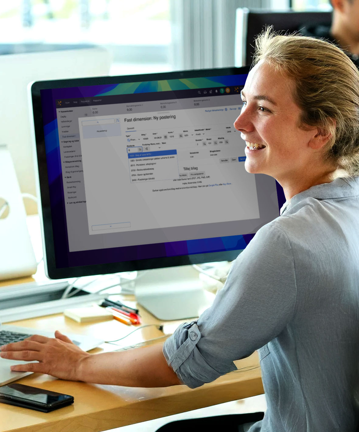 En kvinde sidder ved en computer og smiler til noget eller nogen på venstre side af hende