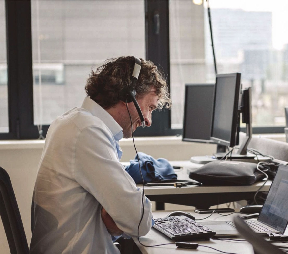 En mand sidder med headset på ved en computer og smiler