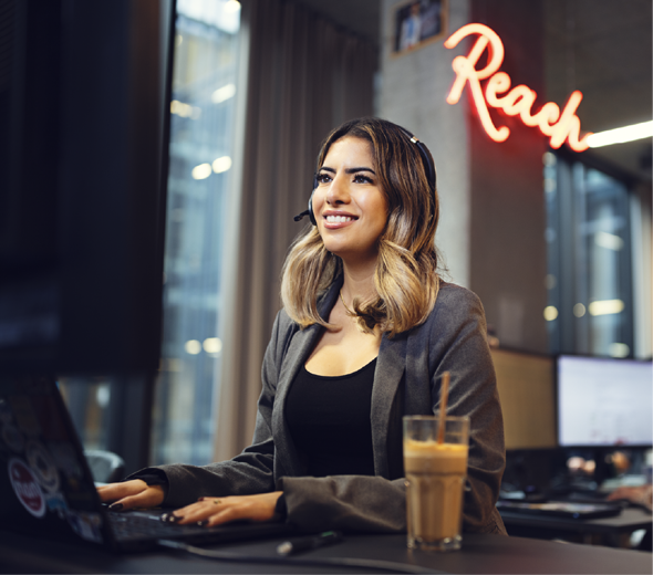 En kvinde med headset på sidder ved en computer og kigger smilende på skærmen