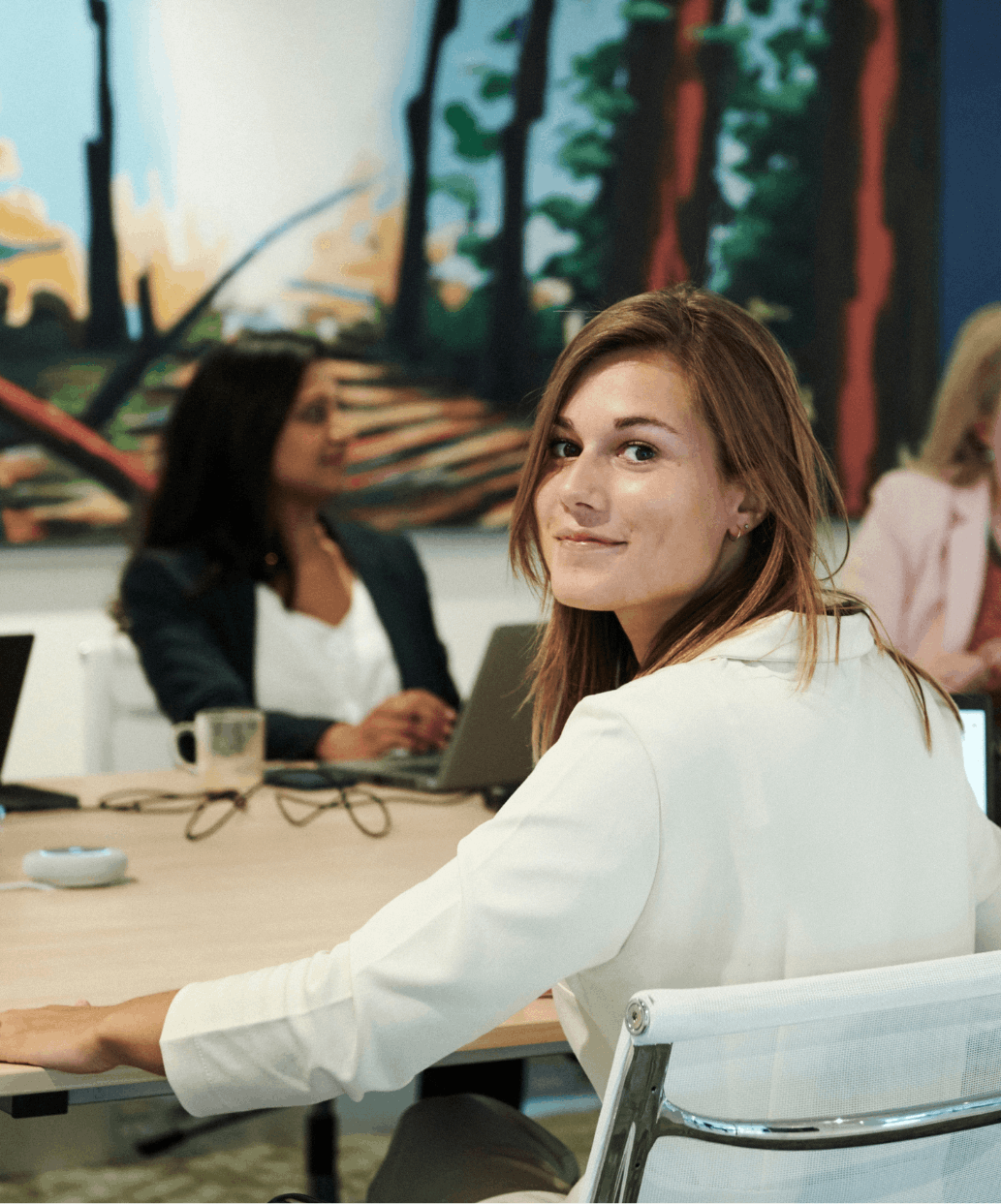 En kvinde i hvid skjorte sidder ved et stort bord og kigger ind i kameraet over skulderen